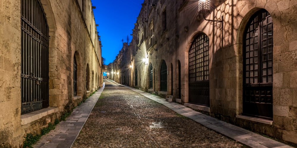 An enchanting view of the historic Knights Street in Rhodes, Greece, as evening falls. The cobblestone path is illuminated by soft lantern light, casting intricate shadows on the ancient stone buildings that line the street