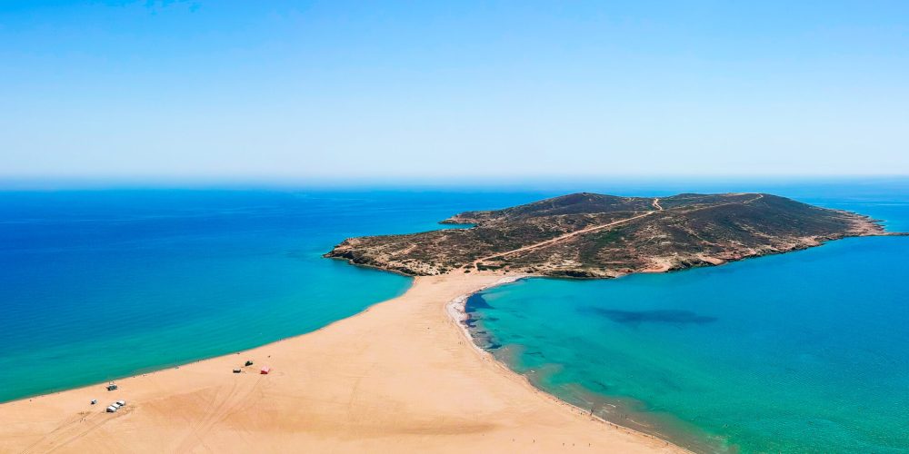 The stunning Prasonisi Beach in Rhodes, Greece, features a unique sandy spit that connects to a small island, creating a picturesque scene with turquoise waters on both sides