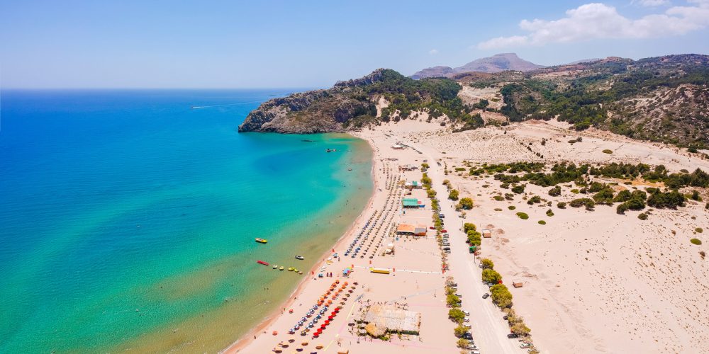 This breathtaking aerial view captures the pristine sandy beach in Rhodes, Greece, lined with rows of vibrant umbrellas and set against the backdrop of crystal-clear turquoise waters and rugged hills