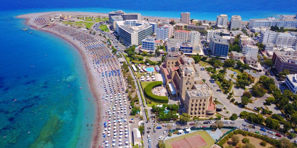 An aerial view of the bustling beach and cityscape in Rhodes, Greece, showcasing a mix of modern buildings, crowded beach umbrellas, and the stunning turquoise waters of the Aegean Sea.